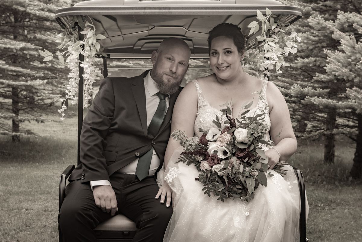 A newlywed couple smiles in a golf cart decorated with foliage, surrounded by a serene wooded setting.