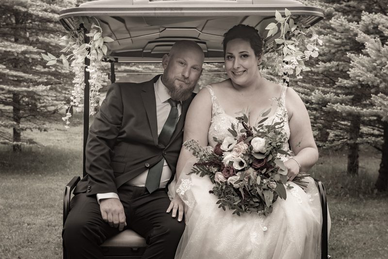 A newlywed couple smiles in a golf cart decorated with foliage, surrounded by a serene wooded setting.