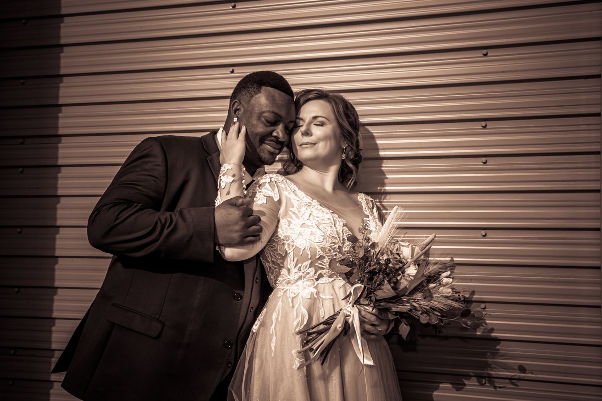 Jennifer and Kevin share a tender embrace against a corrugated metal wall, bathed in warm sunlight that casts long shadows.
