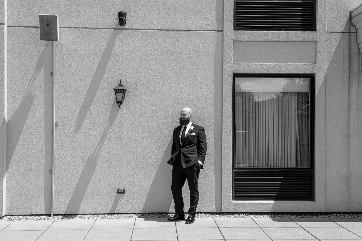 A bearded man in formal wear stands against a stark building wall, casting long shadows in bright sunlight.