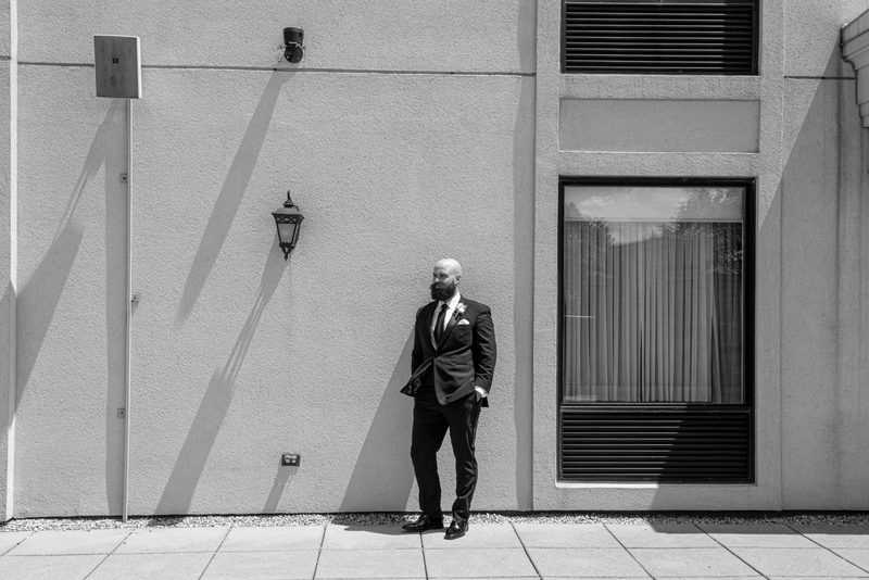 A bearded man in formal wear stands against a stark building wall, casting long shadows in bright sunlight.