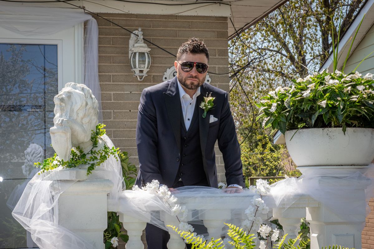 A groom in a dark suit and sunglasses stands on a decorated balcony with floral arrangements and a stone lion statue.