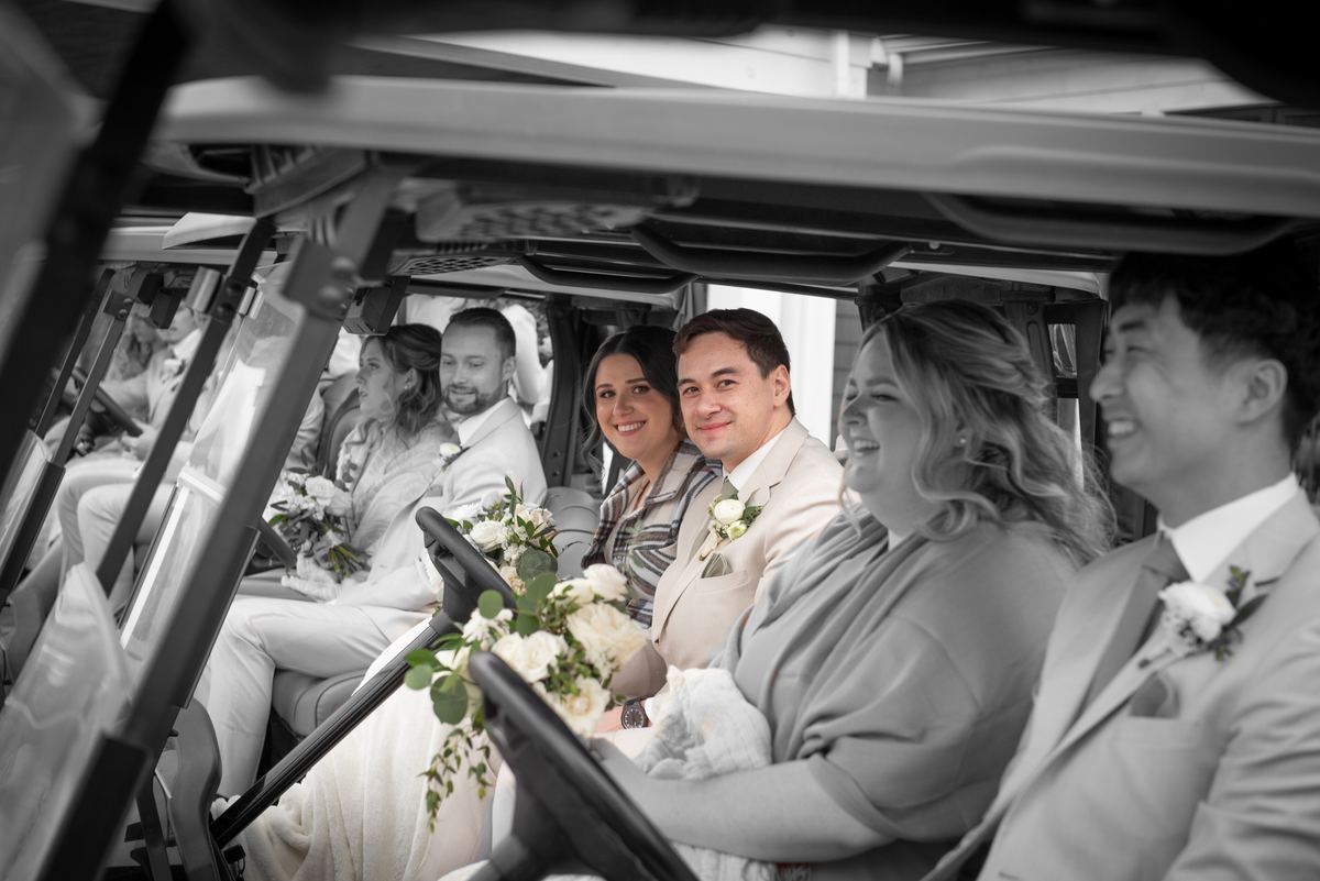A smiling bride and groom with guests in elegant attire ride joyfully in a golf cart, illuminated by soft daylight.