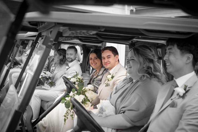 A smiling bride and groom with guests in elegant attire ride joyfully in a golf cart, illuminated by soft daylight.