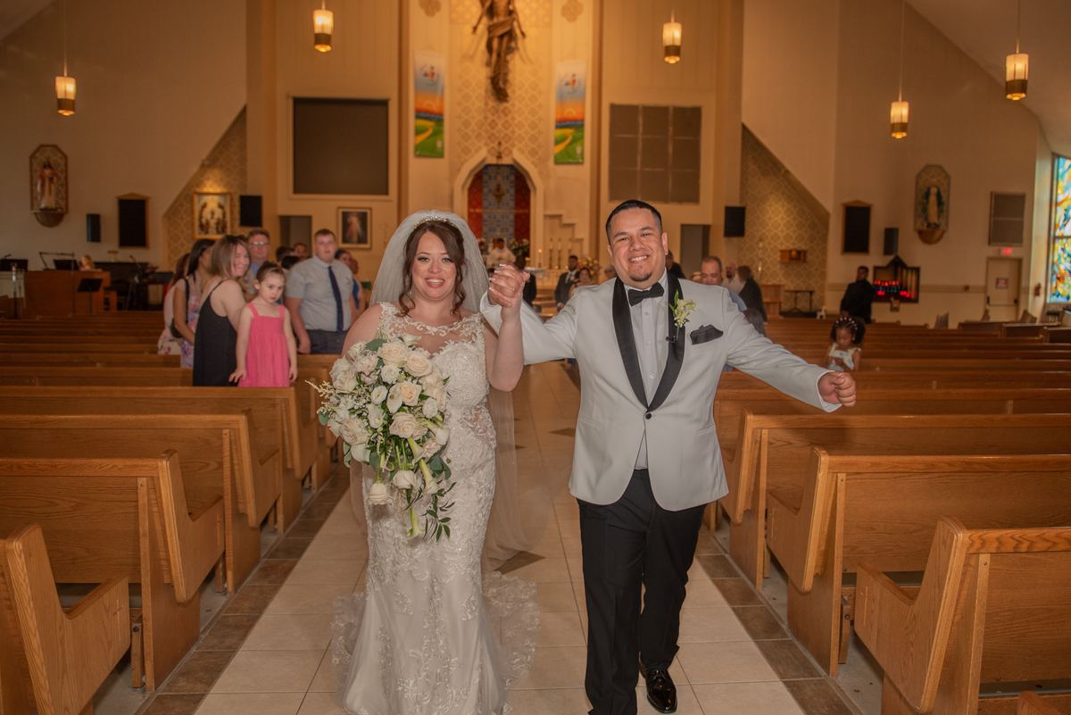 A smiling bride and groom walk down the aisle, hands clasped, surrounded by guests in a warmly lit church.