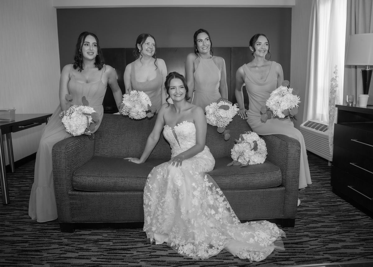 A bride and her bridesmaids sit together on a sofa, smiling with bouquets of flowers in black-and-white tones.