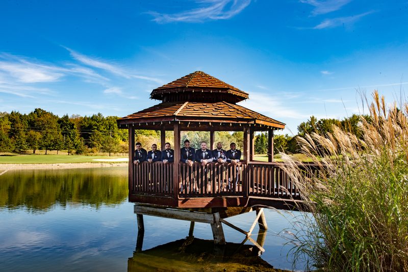 Groomsmen in black tuxedos sit on a wooden gazebo over a calm pond, framed by tall grass and trees, under a bright blue sky.