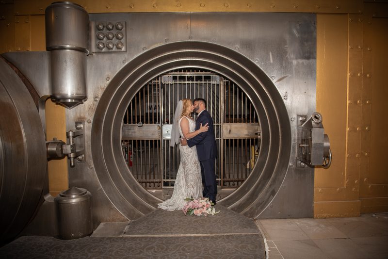 A bride and groom kiss near an industrial vault door, contrasting their attire against the metallic backdrop.