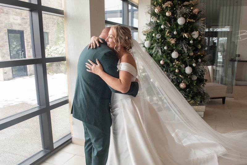 A bride in a white gown embraces her father beside a decorated Christmas tree, with natural light streaming through windows.