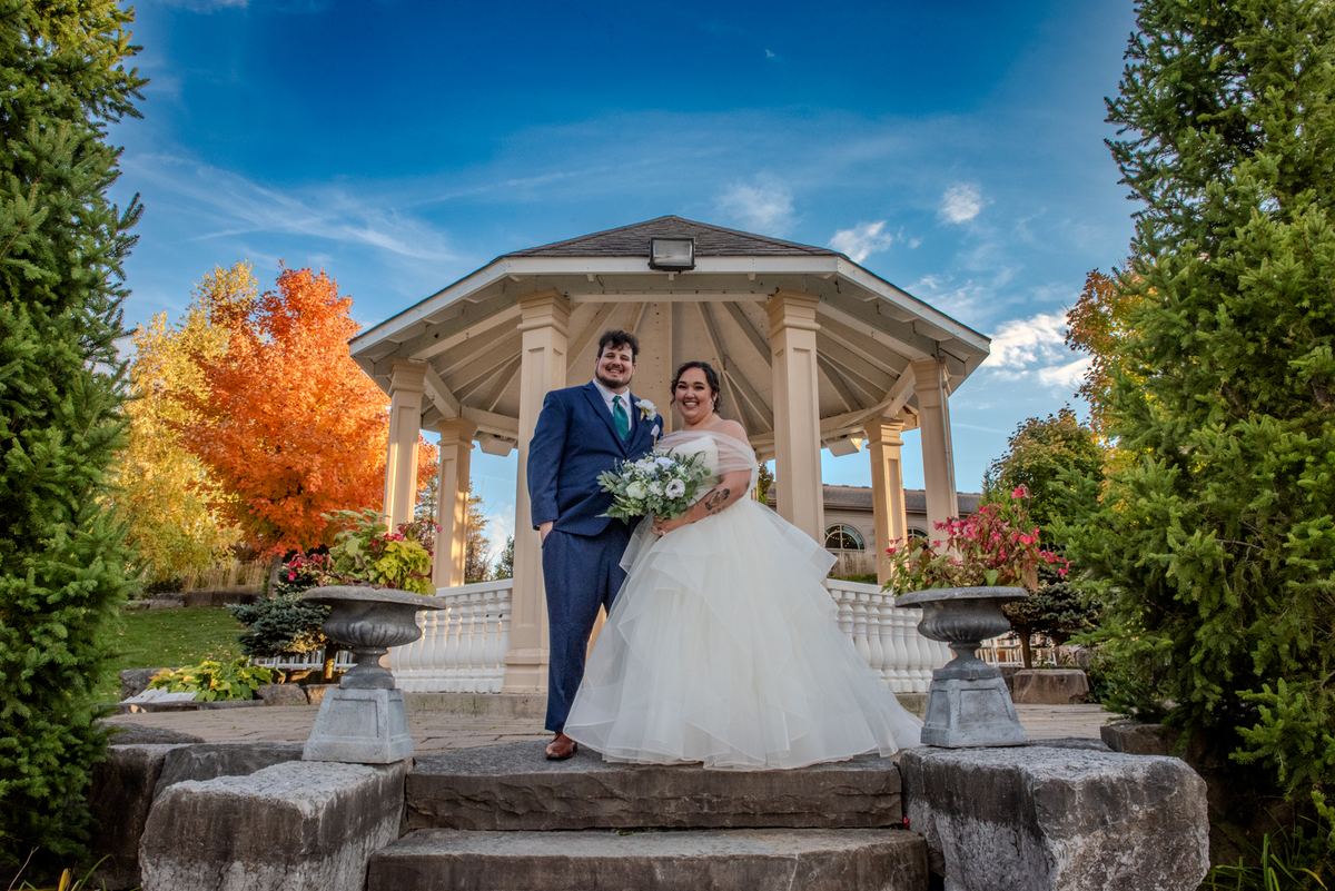 A smiling bride and groom stand on stone steps before a gazebo surrounded by autumn foliage under a clear blue sky.
