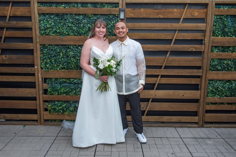 A bride in a white gown and her groom in a barong tagalog stand beside a wooden fence with greenery.