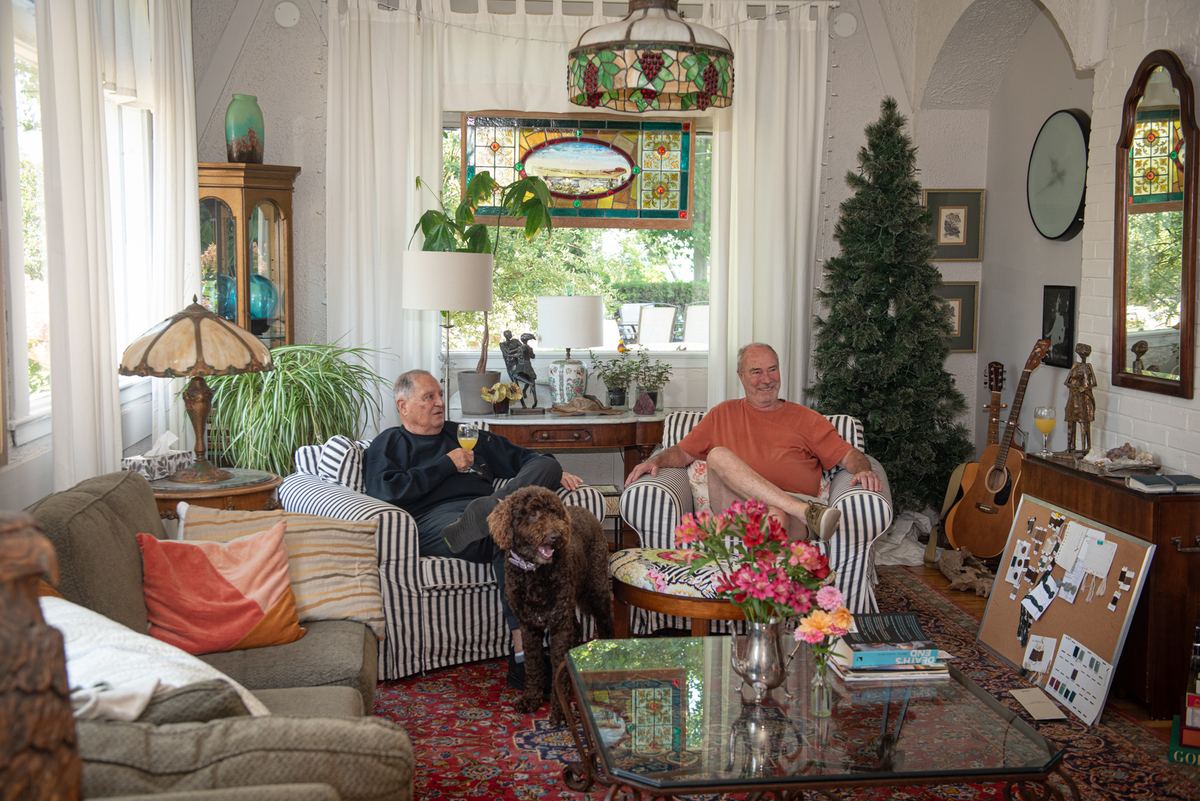 Two men and a dog relax in a cozy living room with natural light and eclectic decor, including a Christmas tree.