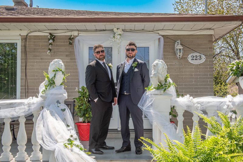 Two men in suits stand on a decorated porch at a same-sex wedding, flanked by veiled statues and greenery.