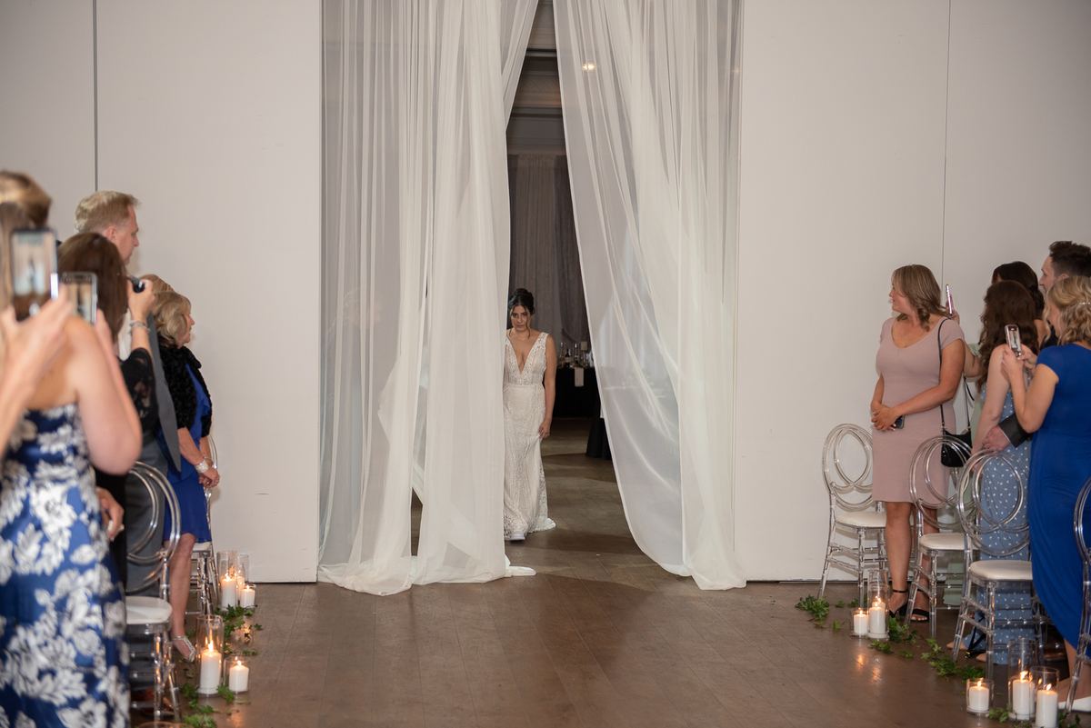 A bride in a white gown stands at the curtain's edge, ready to walk down the candlelit aisle as guests watch on their phones.