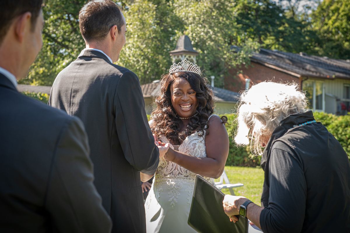 A bride in a white lace gown laughs beside her groom during an outdoor ceremony, illuminated by dappled sunlight.