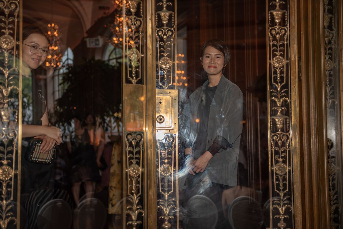 Two women observe through a golden-framed glass door in a sophisticated setting, illuminated by soft chandeliers.
