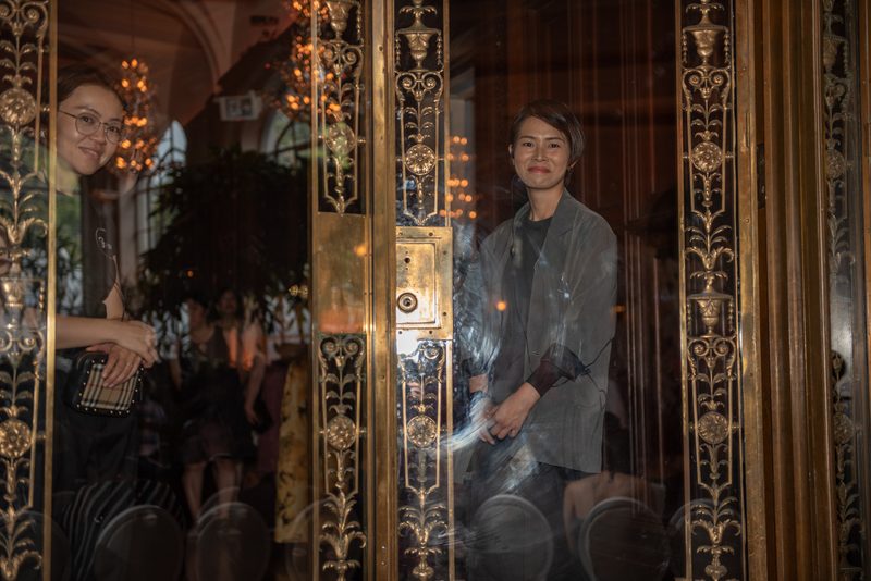 Two women observe through a golden-framed glass door in a sophisticated setting, illuminated by soft chandeliers.