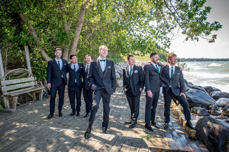 A groom and his groomsmen in dark suits and green ties pose playfully beside a rocky shoreline with trees overhead.
