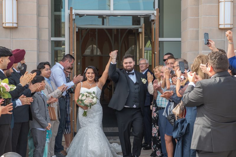 A bride and groom exit a modern church with arms raised as guests clap and capture the moment on phones.