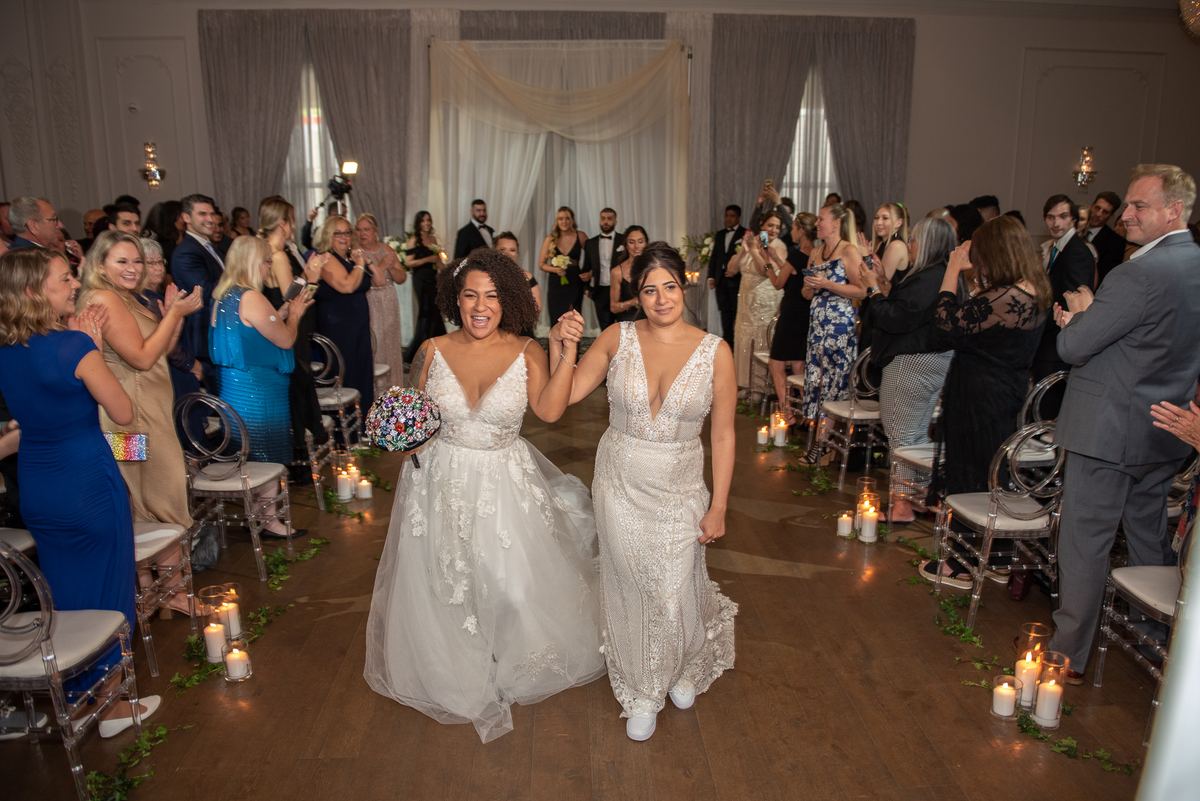 Two brides in white gowns walk down a candlelit aisle decorated with greenery, smiling as guests applaud.
