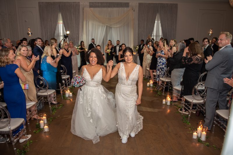 Two brides in white gowns walk down a candlelit aisle decorated with greenery, smiling as guests applaud.