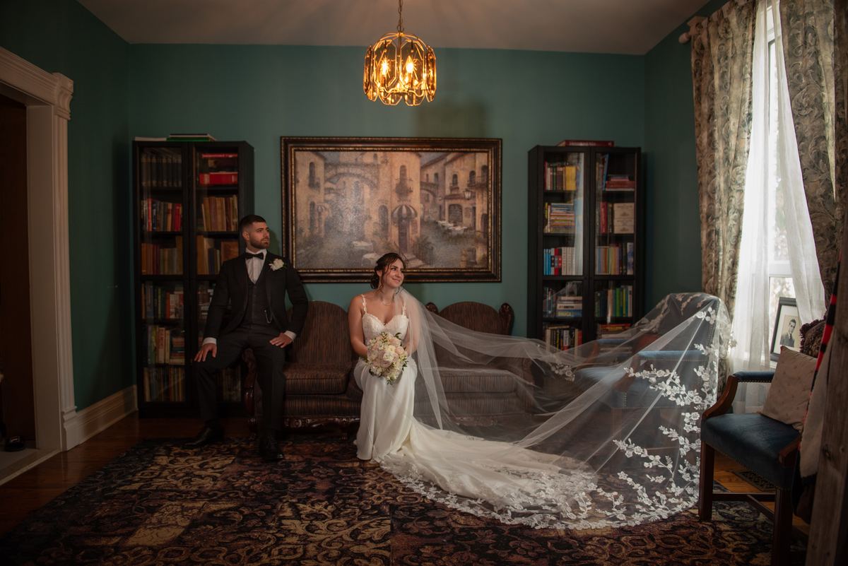 Bride and groom sit together in a warmly lit room with bookshelves, chandelier, and natural light from a window.