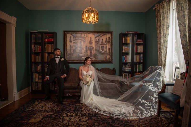 Bride and groom sit together in a warmly lit room with bookshelves, chandelier, and natural light from a window.