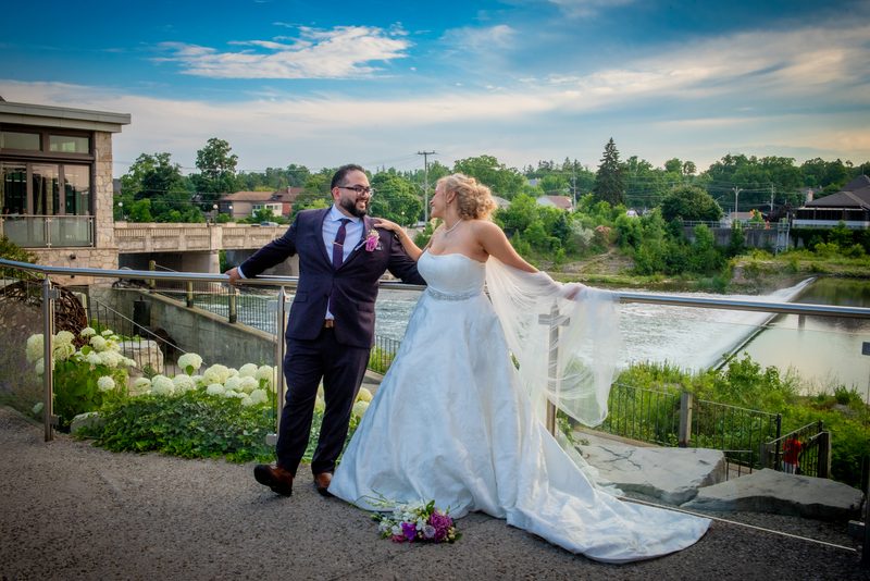 A laughing bride and groom embrace at a riverside overlook, surrounded by lush greenery and a rushing river.