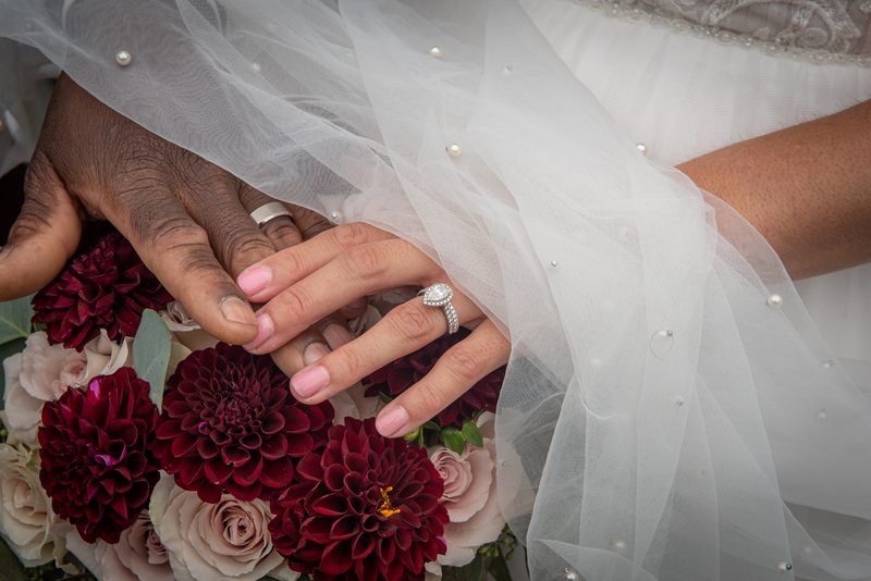 Two hands, one darker and one lighter, holding a bouquet of dahlias and roses with a diamond ring visible.
