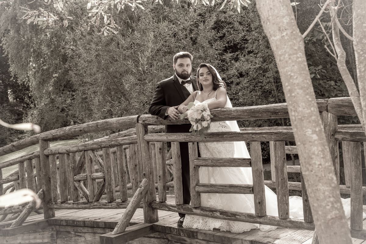 A bride and groom stand on a rustic wooden bridge, framed by trees in soft light, gazing at the camera.