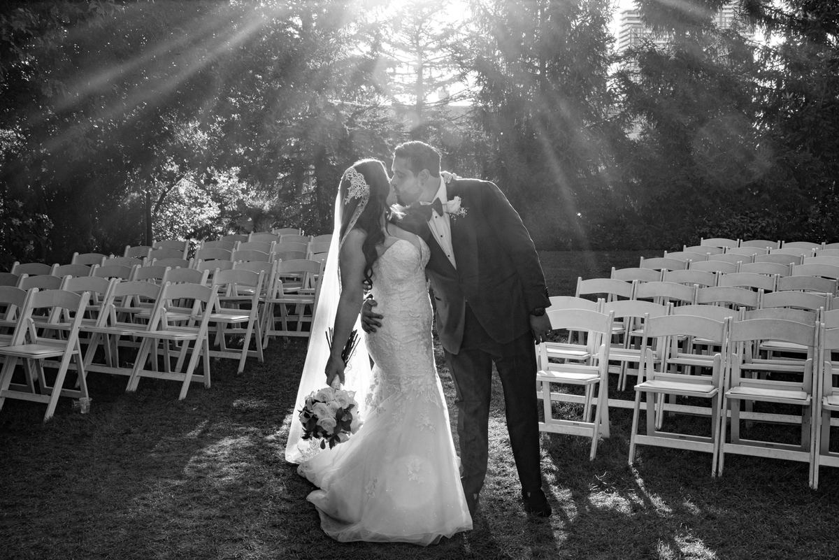 A bride and groom kiss in sunlight, surrounded by white chairs and foliage, with golden rays shining through the trees.