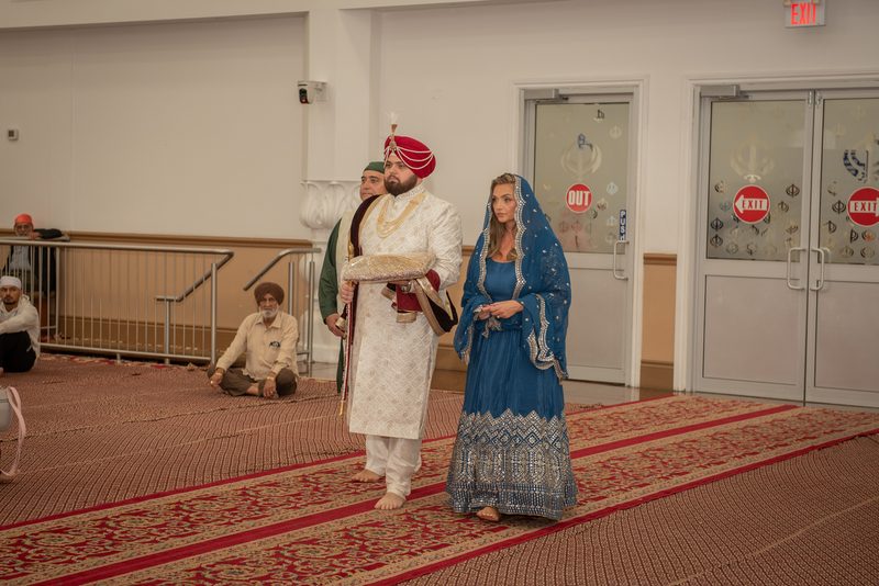A Sikh groom in red turban and white robe walks barefoot beside bride in blue lehenga along patterned carpet inside hall.