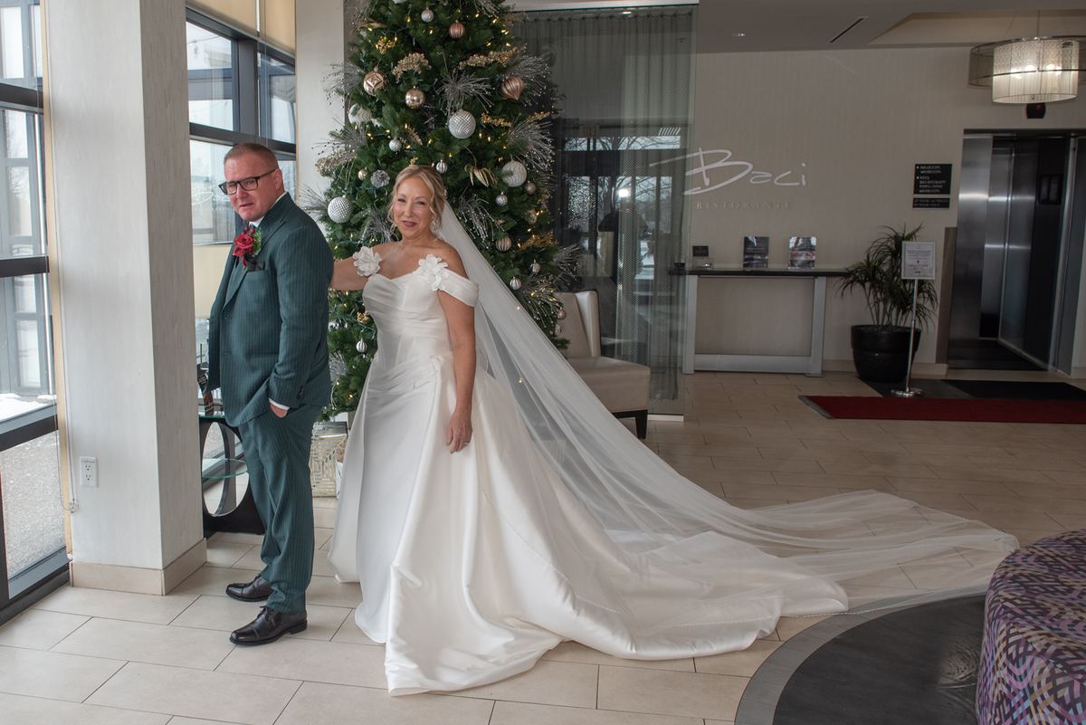A bride in white and groom in teal stand beside a Christmas tree in a hotel lobby, bathed in natural light.