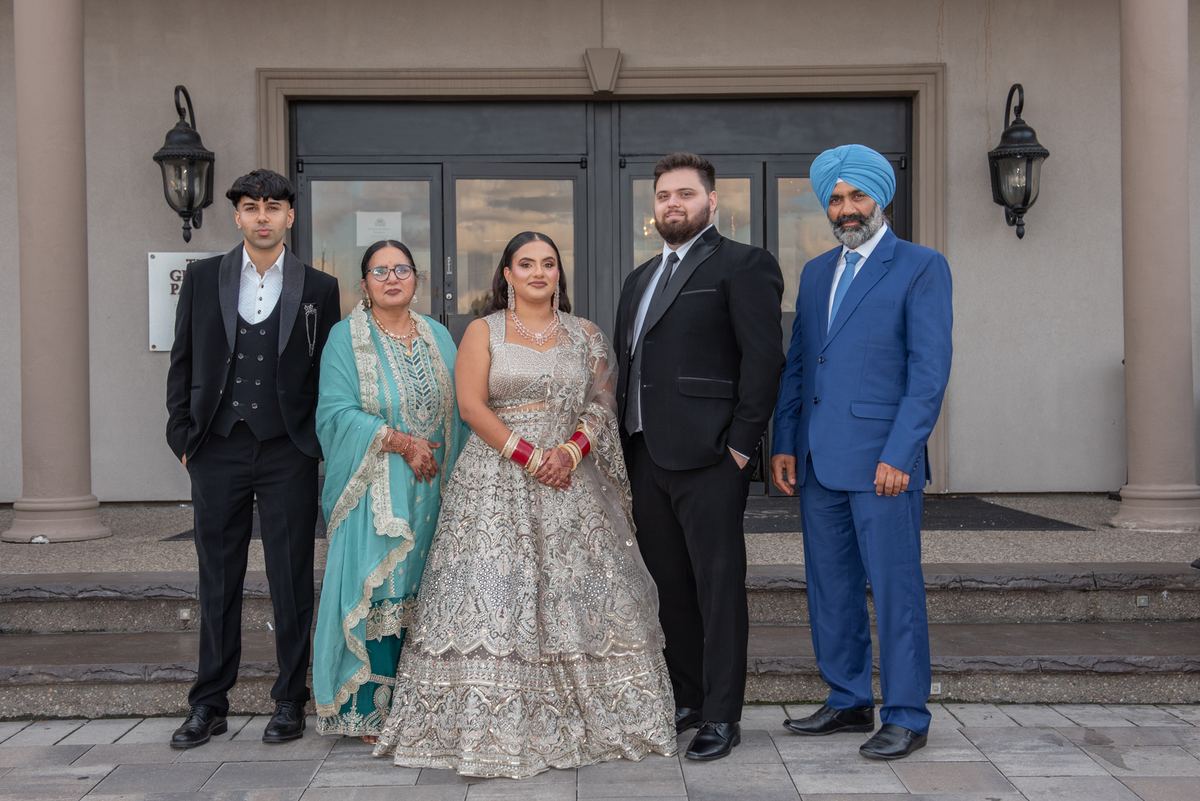 A bride in a silver lehenga and groom in a blue sherwani stand with family members on stone steps before a modern building.