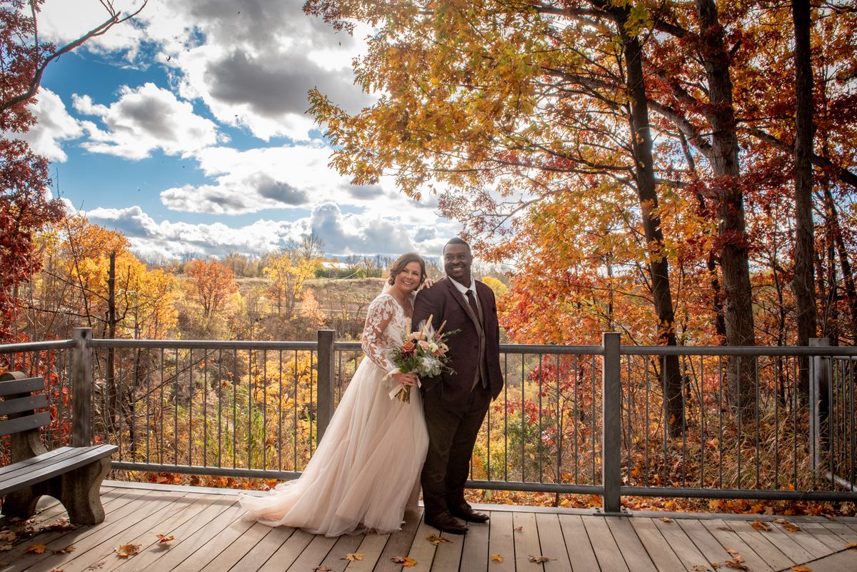 A newlywed couple in formal attire stands on a wooden deck surrounded by autumn foliage and smiling at the camera.