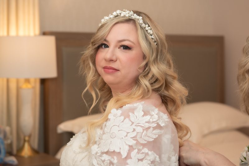 A bride with golden curls and a pearl headpiece looks at the camera as her gown is adjusted in warm hotel room light.