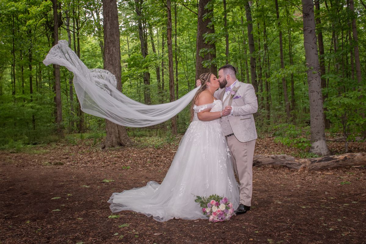 A bride and groom kiss in a sunlit forest, their hands clasped, with her veil fluttering in the breeze.
