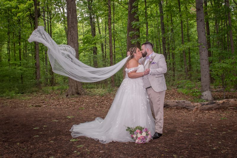 A bride and groom kiss in a sunlit forest, their hands clasped, with her veil fluttering in the breeze.