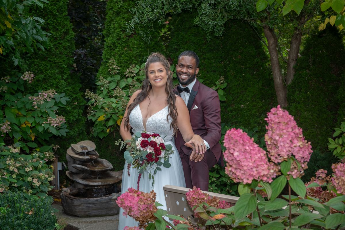 A smiling bride and groom hold hands in a garden with diffused sunlight filtering through the trees.