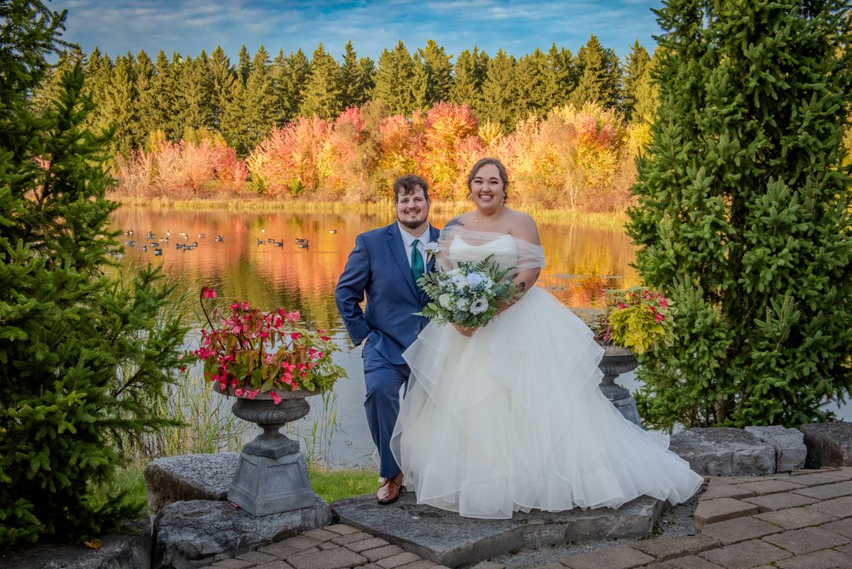 A bride and groom smile by a lake surrounded by autumn leaves and evergreens, with ducks swimming in the background.