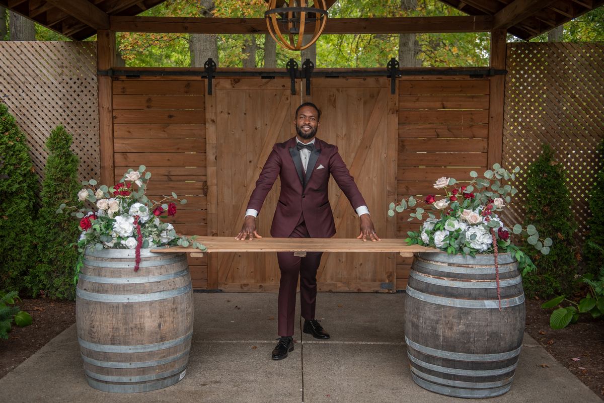 A groom in a burgundy tuxedo stands between two large wine barrels, holding a wooden plank under a rustic pergola.