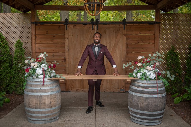 A groom in a burgundy tuxedo stands between two large wine barrels, holding a wooden plank under a rustic pergola.