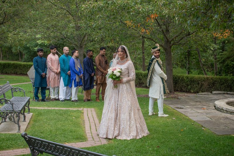 A bride in a cream lehenga walks toward the groom in a white sherwani along a garden path lined with groomsmen.