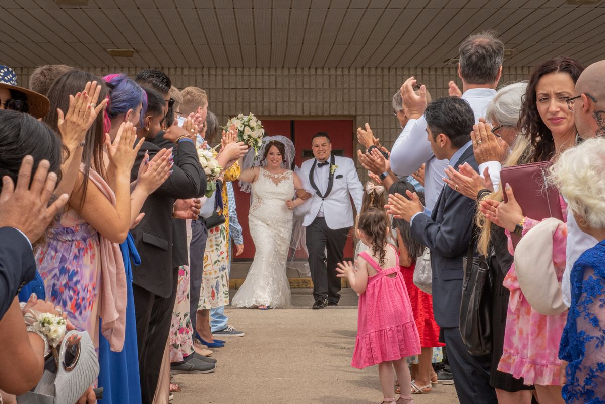 A bride in white lace and groom in white tux walk down the aisle toward cheering guests, surrounded by family and friends.