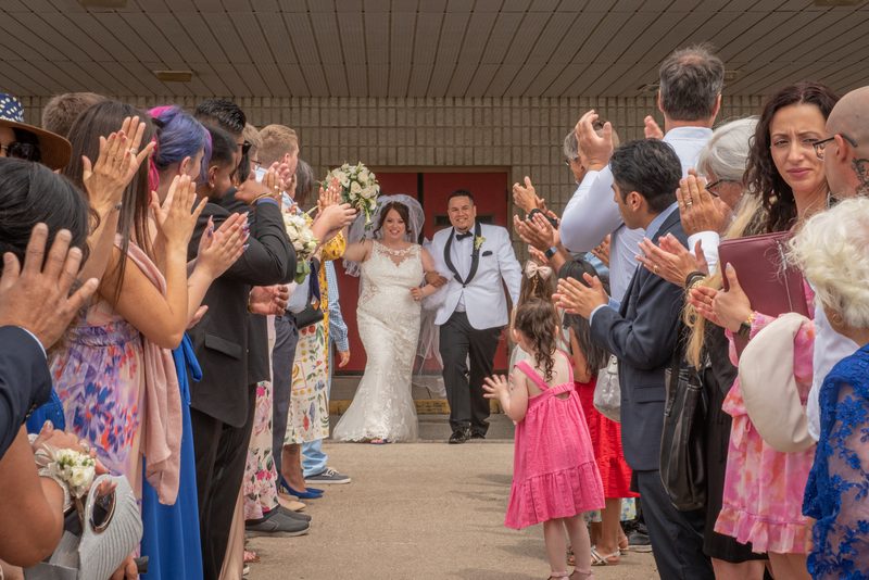 A bride in white lace and groom in white tux walk down the aisle toward cheering guests, surrounded by family and friends.