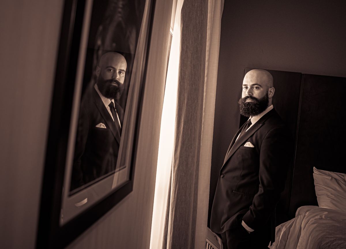 A man in a suit stands thoughtfully in a dim hotel room, with light streaming through a curtain beside him.