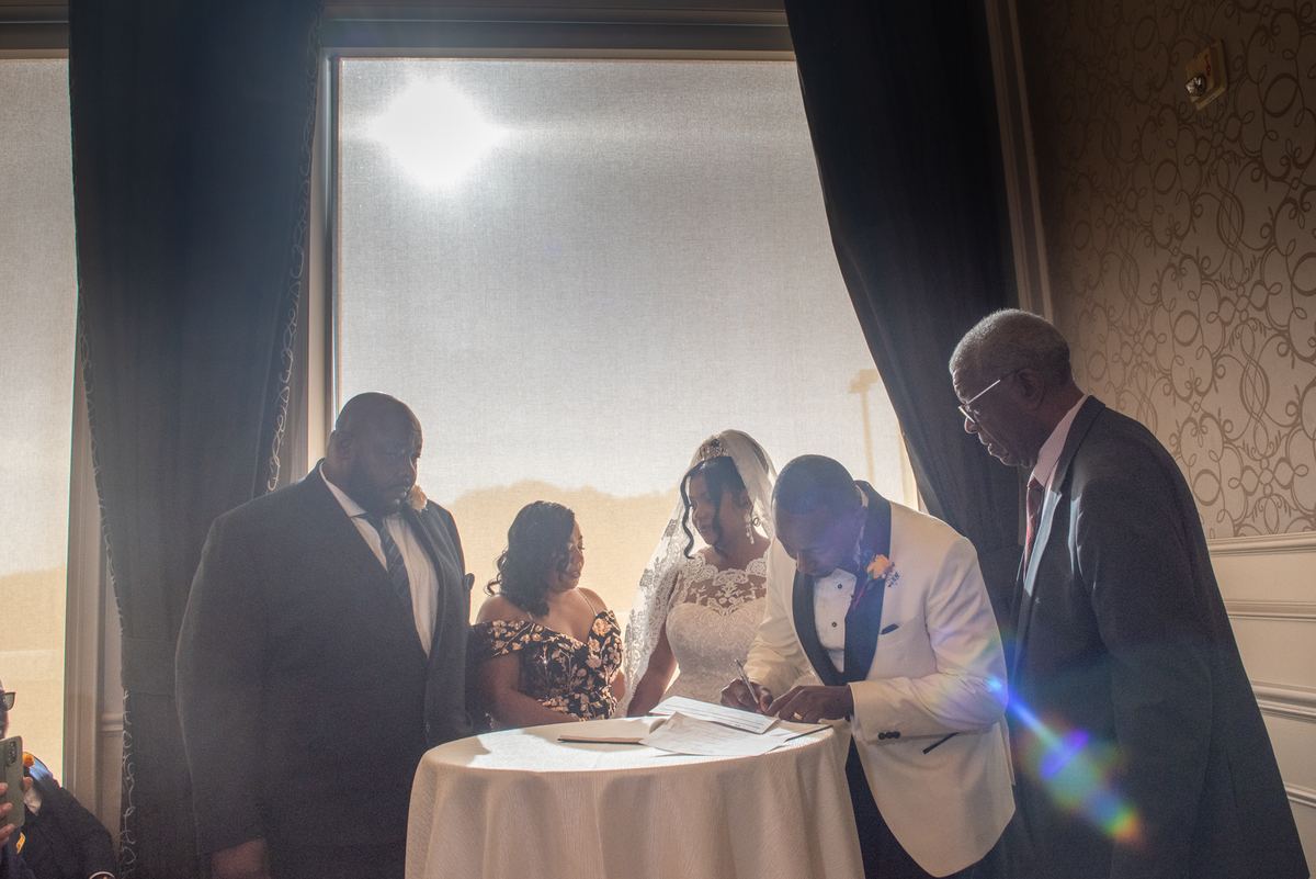 Bride and groom signing documents beside a table with sunlight creating lens flare, guests watching.