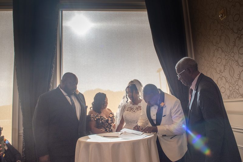 Bride and groom signing documents beside a table with sunlight creating lens flare, guests watching.