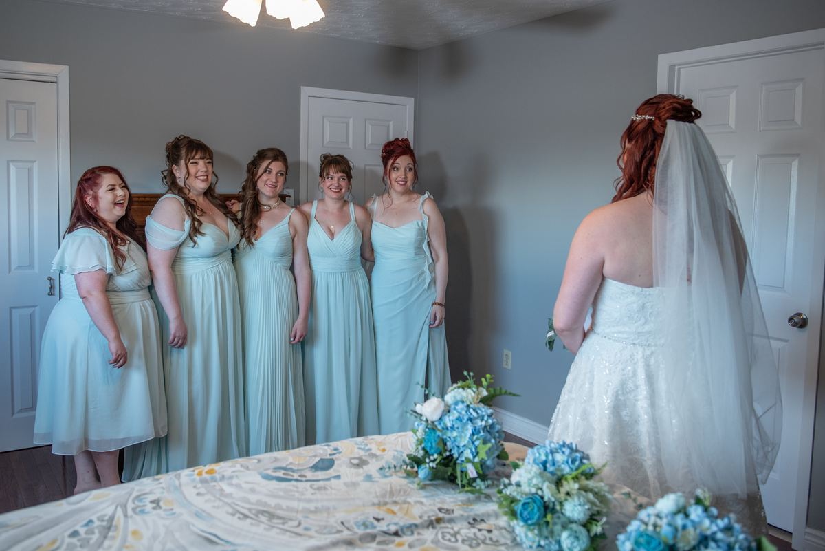 A bride in a white gown faces her laughing bridesmaids in light blue dresses near a table with blue and white flowers.