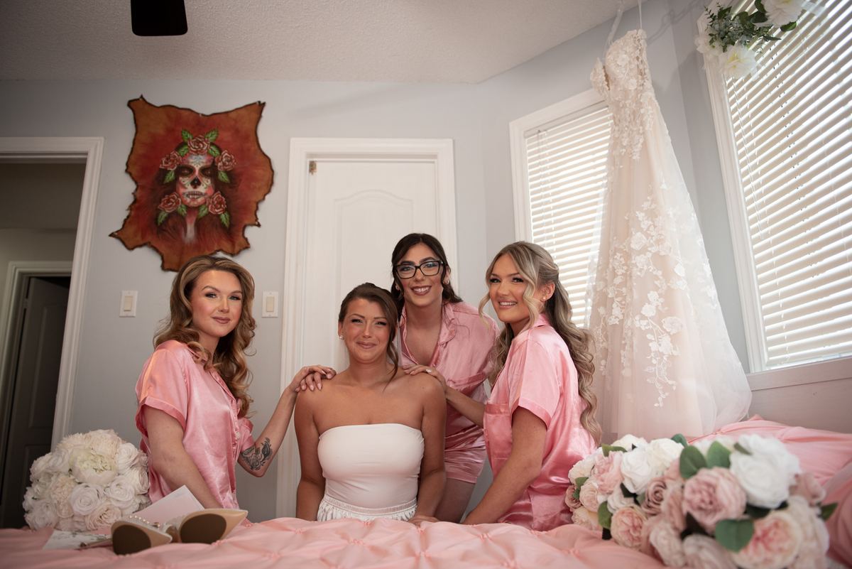 A bride in a white gown and four bridesmaids in pink robes sit on a bed surrounded by roses and a hanging wedding dress.